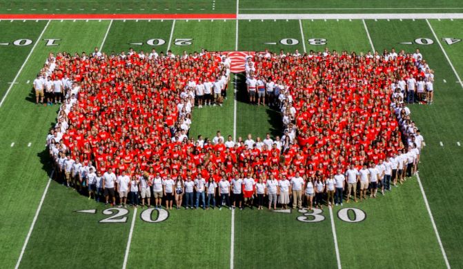 Large group of students stand in red shirts on the University of Utah football field in the shape of the Block U.