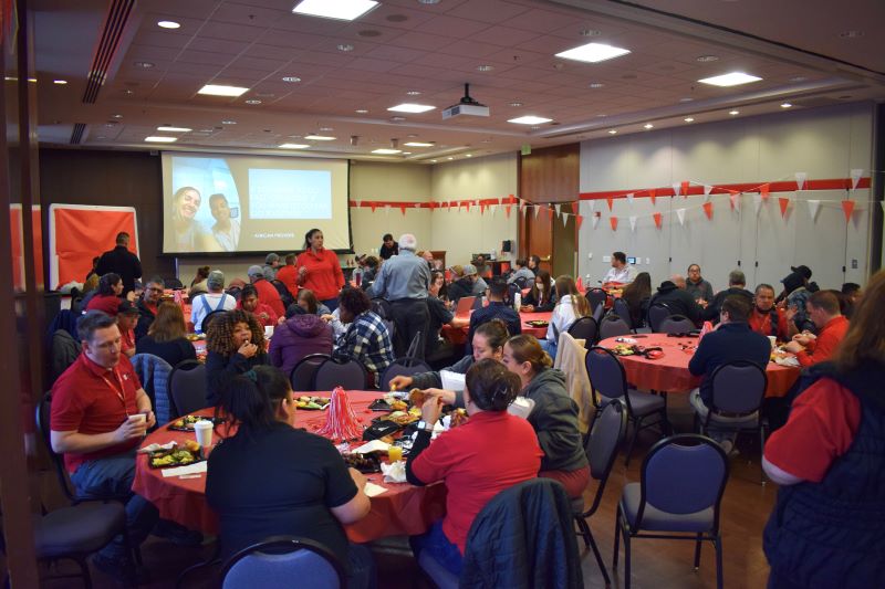 photo of a large room full of people seated at round tables with red table cloths while a slide show plays on a large projector screen at the front of the room.