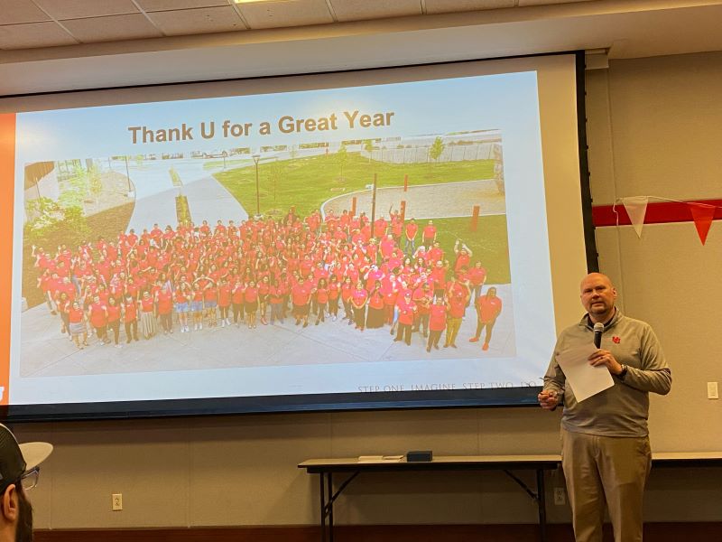 HRE Staff member stands speaking into microphone in front of a slide which reads, "Thank U for a Great Year!"