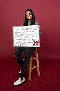 Person sits on a wooden bar stool holding a whiteboard which reads, "I am an empowered and proud Indian American Woman. I am a Multi-Lingual College Student with a double-major. By running an international non-profit, I stand with equality and authenticity. #BeYou #LoveUrself."