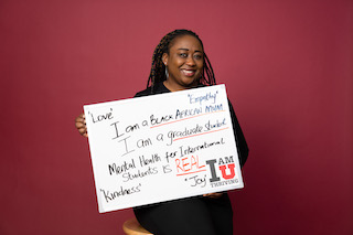 Person holds a whiteboard which reads, "Love, Empathy, I am a Black African Mum. I am a Graduate Student. Mental Health for International Students is REAL. Kindness. Joy."