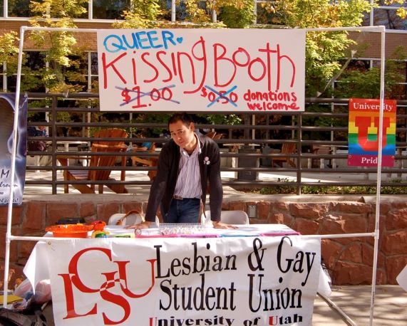 student stands at a "Queer Kissing Booth" table with a sign for the Lesbian & Gay Student Union