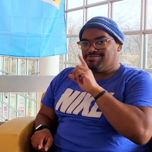 Photo of LJ Williams wearing a blue t-shirt and blue hat, sitting in a chair and using sign language to speak with his advisor.