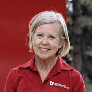 Headshot of Ann House with a red background. She is wearing a red collared shirt. 