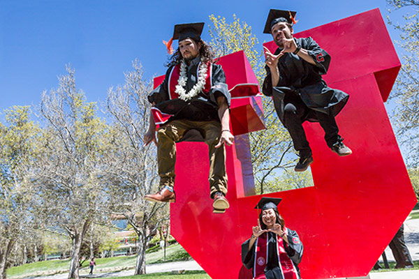 students wearing graduation caps and gowns are captured mid-air jumping off block U.